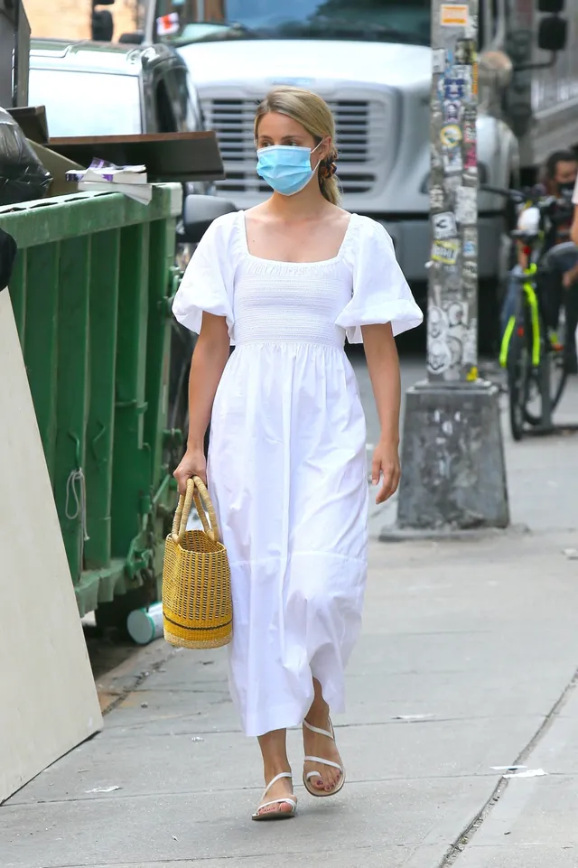 Dianna Agron walking beside a fence in a white dress and flat sandals.