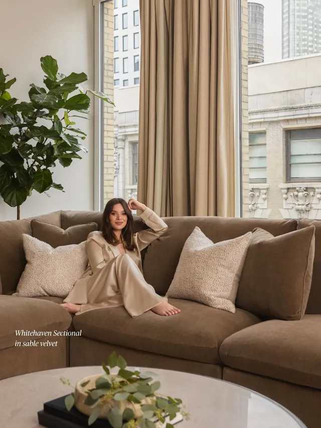 Hailee Steinfeld sitting barefoot on a sofa in a beige outfit.