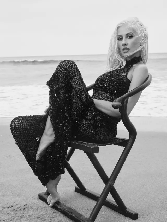 Christina Aguilera sitting by the sea in a dark gown and metallic heels.