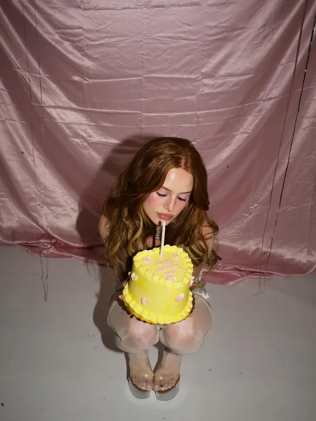 Madelaine Petsch crouching in socks and gold heels while holding a yellow cake.