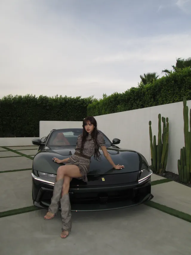 Lisa (Lalisa Manobal) sitting on a vehicle bonnet in tall open-toe heels and a patterned dress.