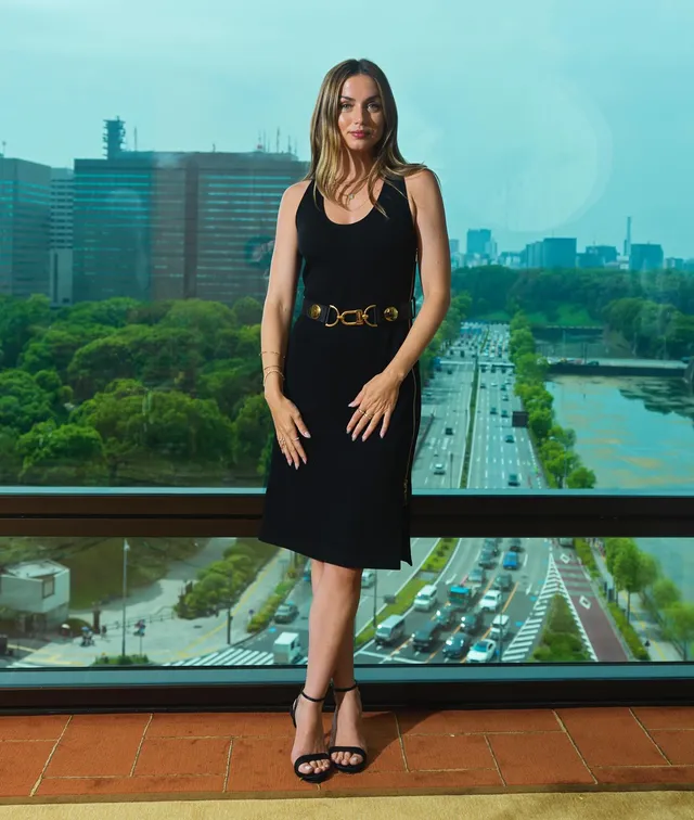 Ana de Armas standing by a city-view window in a black dress and strappy heels.