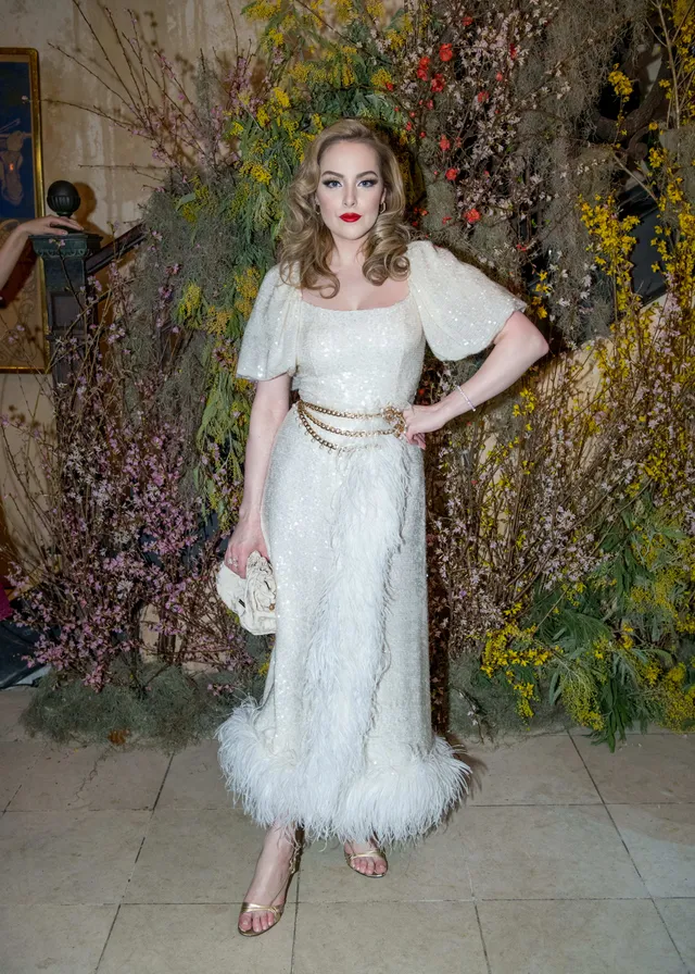 Elizabeth Gillies standing at an event in a white feather-trimmed dress.
