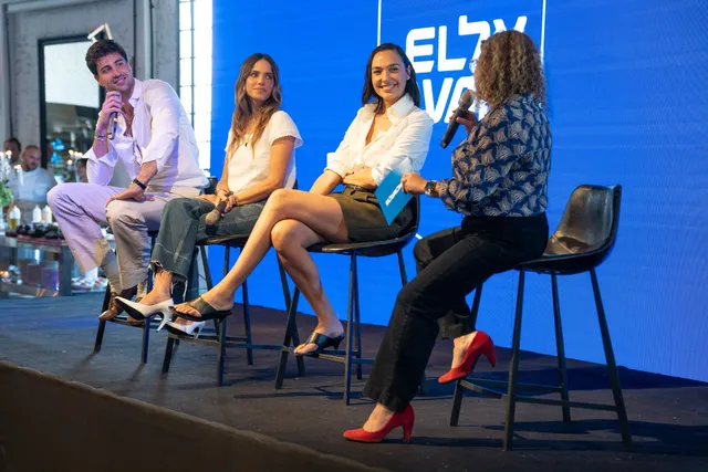 Gal Gadot sitting on a panel in a pale outfit and heels.