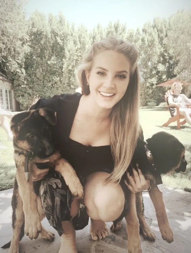 Lana Del Rey crouching beside dogs in an outdoor photograph.