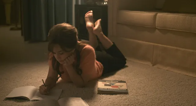 Cailee Spaeny lying barefoot on a bedroom carpet beside open books.