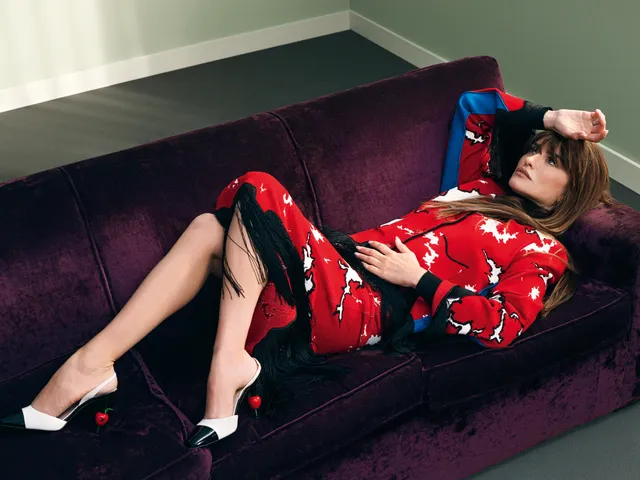 Penélope Cruz reclining on a purple sofa in a red patterned dress and heels.