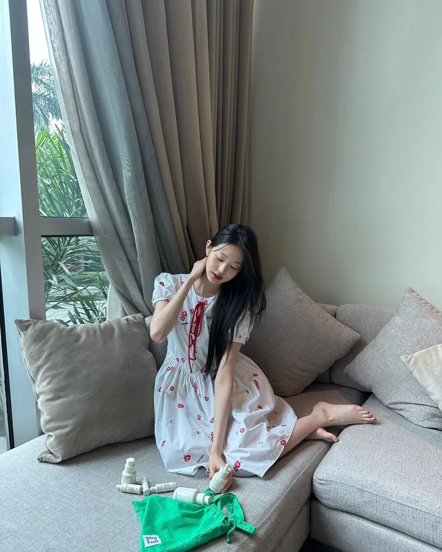 Jang Wonyoung sitting barefoot on a sofa by a window.