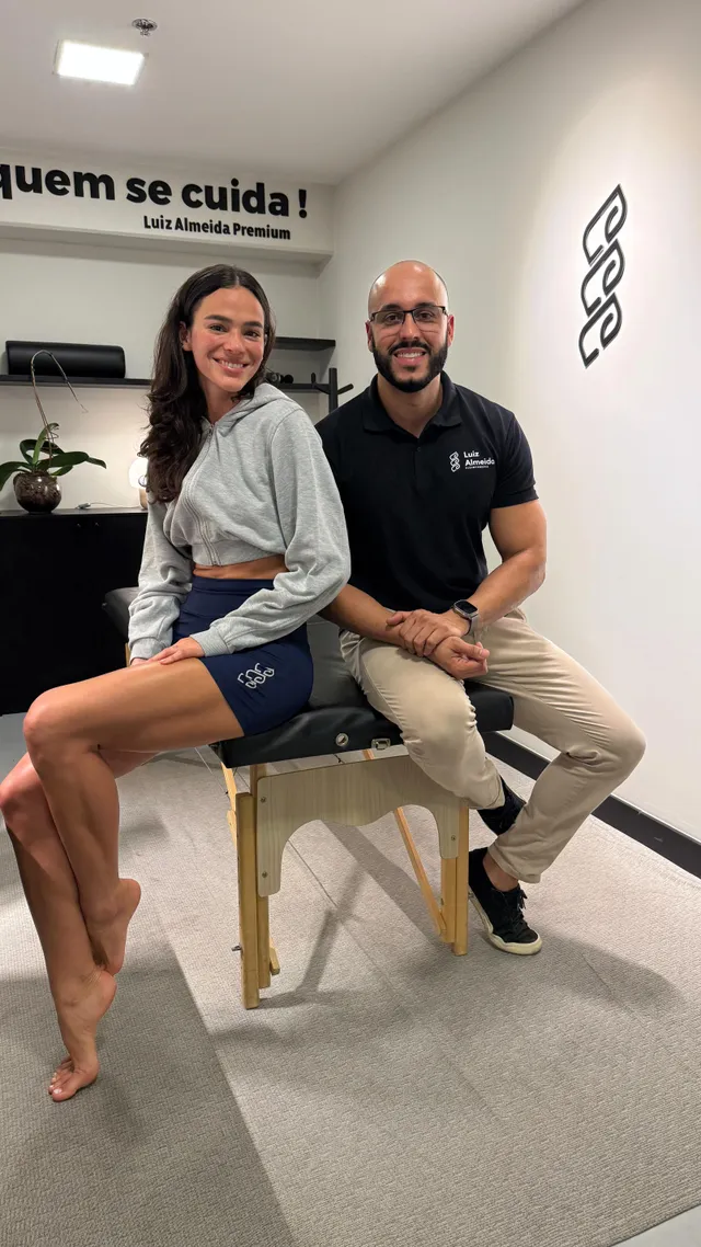 Bruna Marquezine sitting barefoot beside a man indoors in a grey top and shorts.