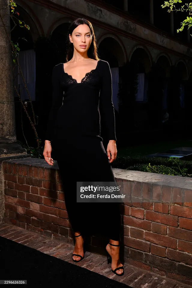 Irina Shayk standing outdoors in a black dress and heeled sandals.
