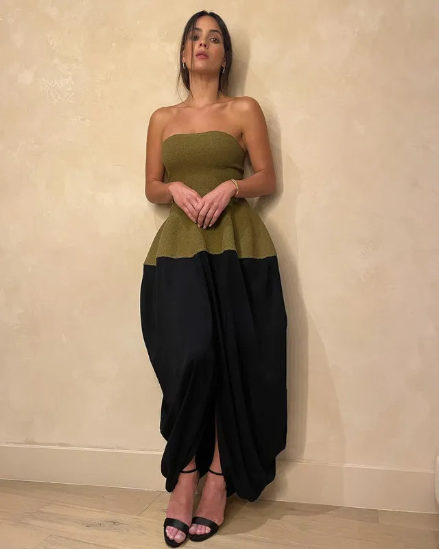Adria Arjona standing in an olive top, black trousers and black heeled sandals.