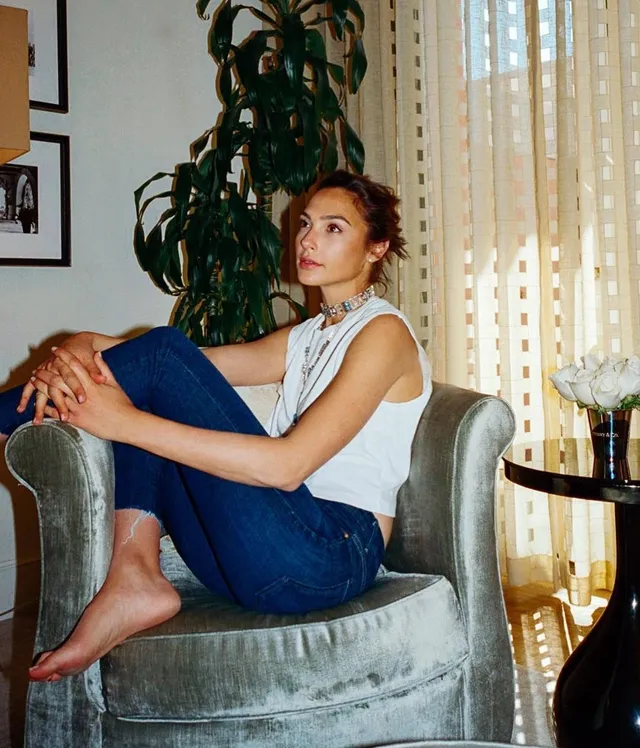 Gal Gadot sitting barefoot in a pale armchair wearing jeans.