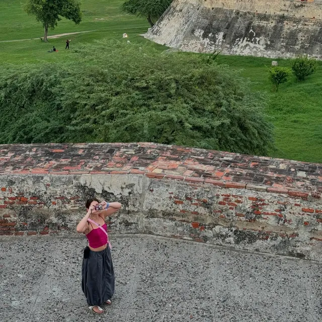 Nicole Wallace standing outdoors beside a stone wall in a pink top and long dark skirt.