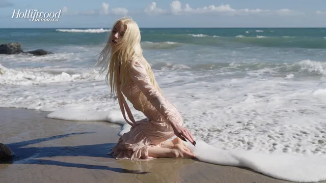 Anya Taylor-Joy kneeling barefoot on a beach near the surf.