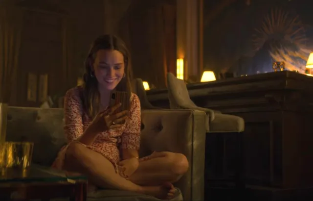 Victoria Pedretti sitting cross-legged on a sofa during a screen scene.