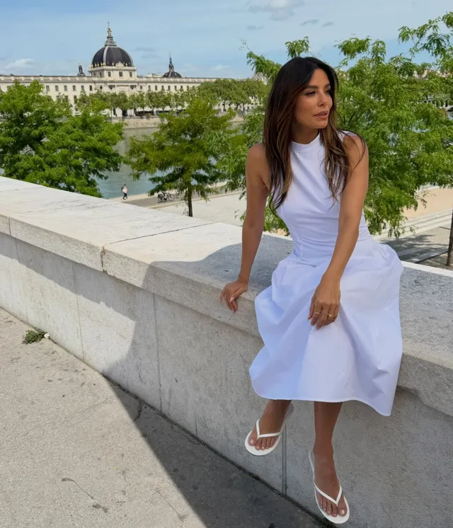 Eva Longoria sitting on a stone wall in a white dress and flat sandals.
