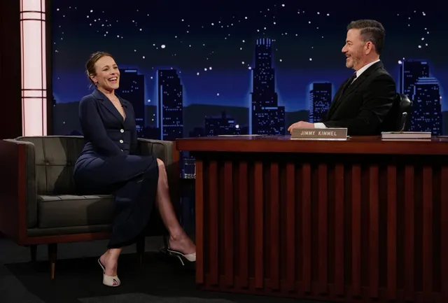 Rachel McAdams sitting during a television interview in a dark outfit and pale heels.
