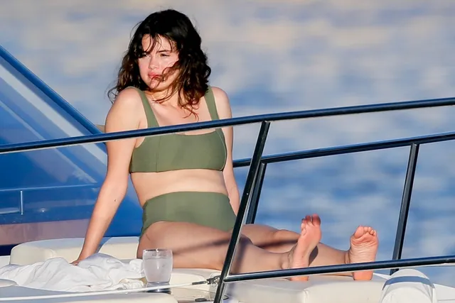 Selena Gomez: seated barefoot on a boat in a green swimsuit.