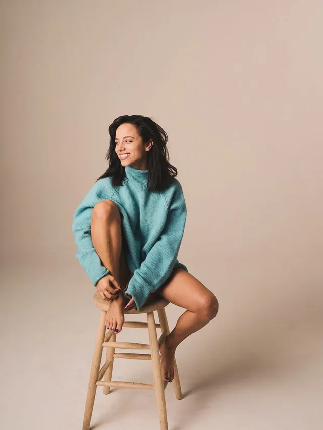 Kiana Madeira sitting barefoot on a wooden stool in a teal sweater.