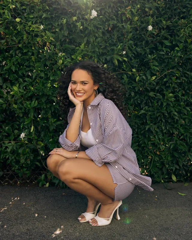 Madison Pettis crouching outdoors in a striped shirt and white heels.