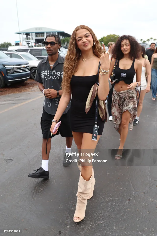 Madison Bailey walking outdoors in a black mini dress and pale boots.