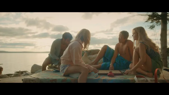 Emily Alyn Lind sitting barefoot with friends beside the sea at sunset.
