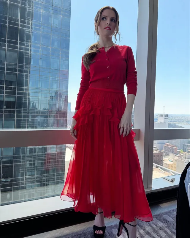Anna Kendrick standing beside a window in a red dress and black heels.