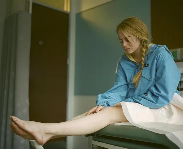 Emma Stone sitting in a blue shirt with her bare legs and feet extended.