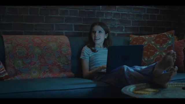 Anna Kendrick reclining on a sofa with a laptop and bare soles visible.
