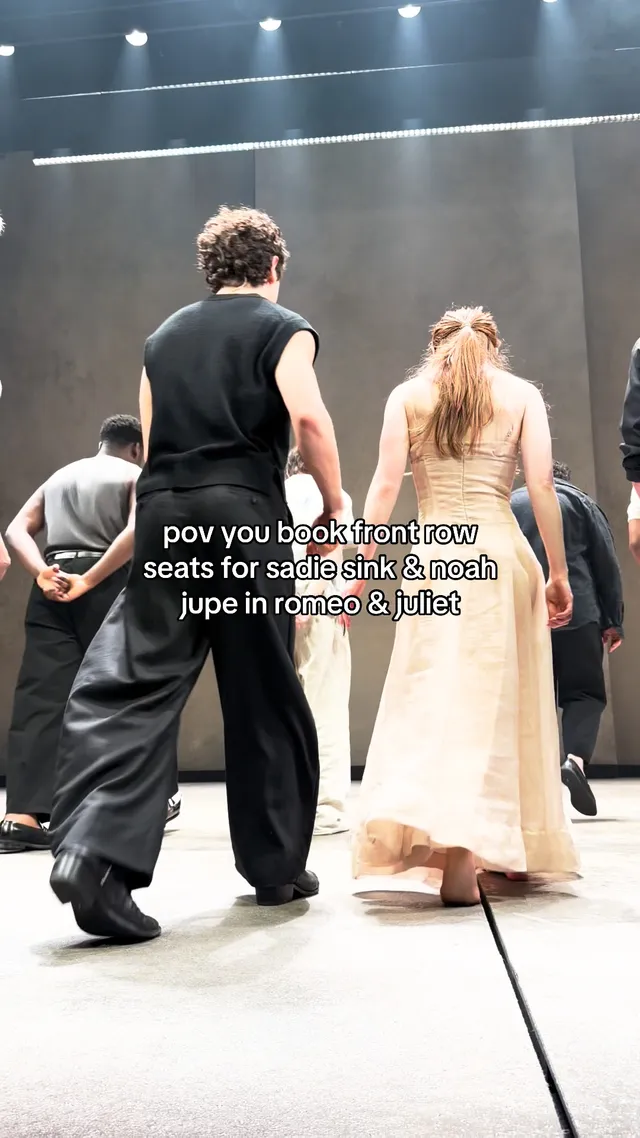 Sadie Sink standing barefoot on a theatre stage in a pale dress.