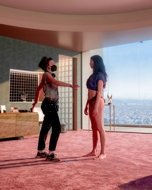 Margaret Qualley a standing screen scene in a pink room beside a companion.