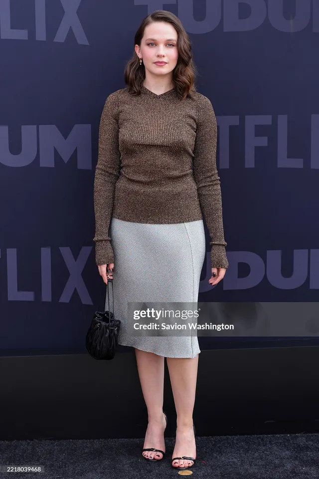 Cailee Spaeny standing at a film event in a grey skirt and black heels.