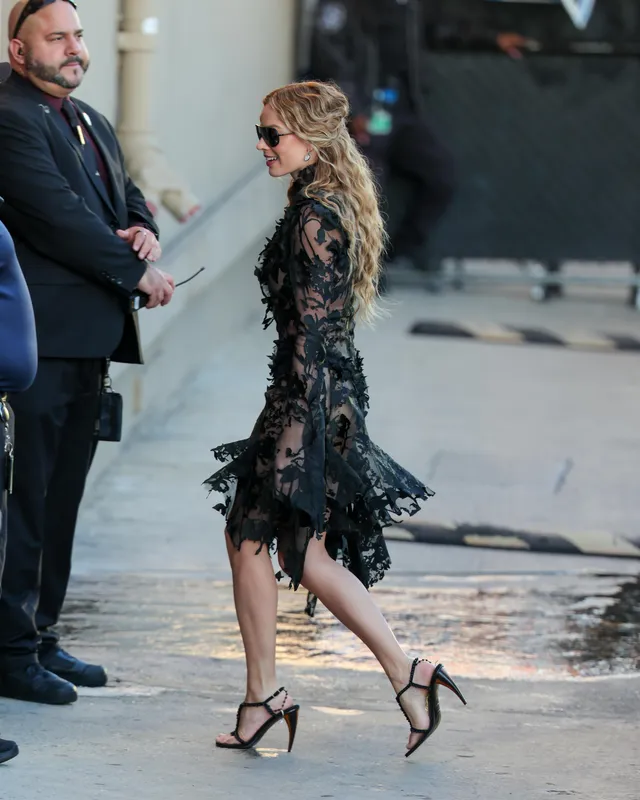 Margot Robbie walking outside in a black lace dress and heels.