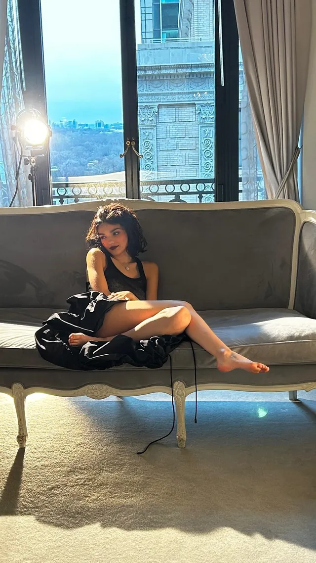 Rachel Zegler sitting barefoot on a sofa beside a tall window.