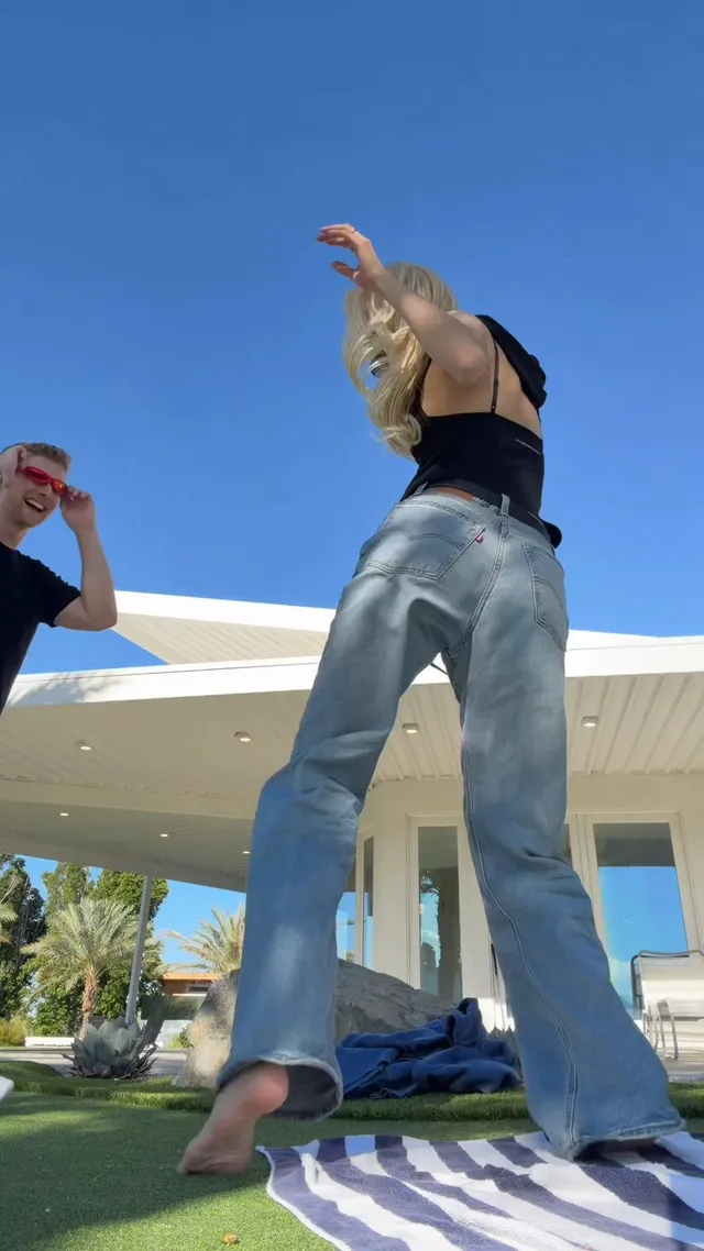 Rosé dancing barefoot on a lawn in a dark sleeveless top and blue jeans.