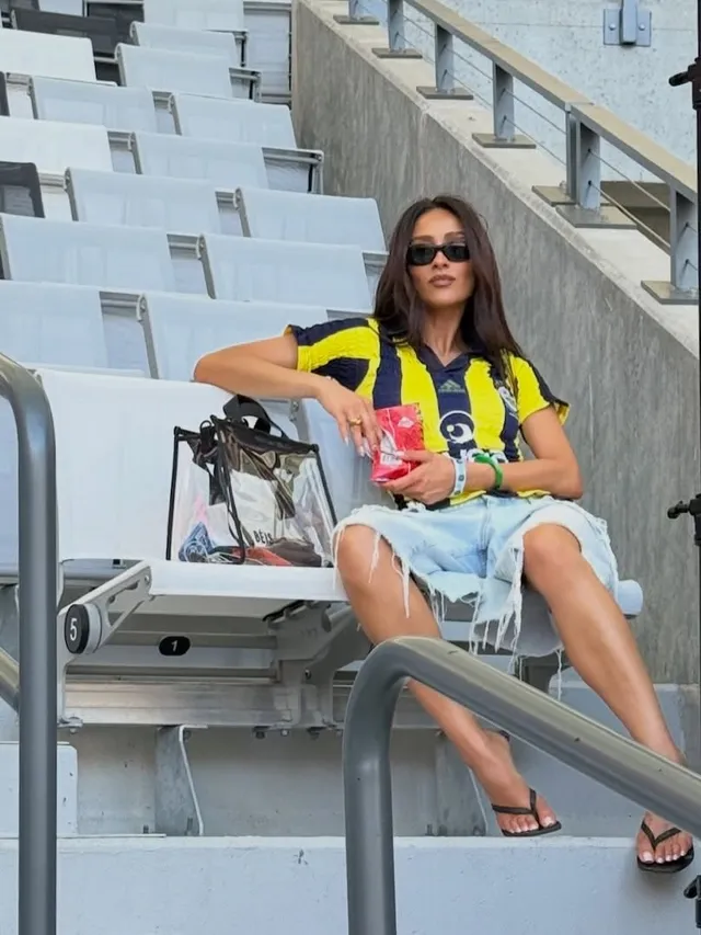 Shay Mitchell sitting in stadium seats in denim shorts and flip-flops.