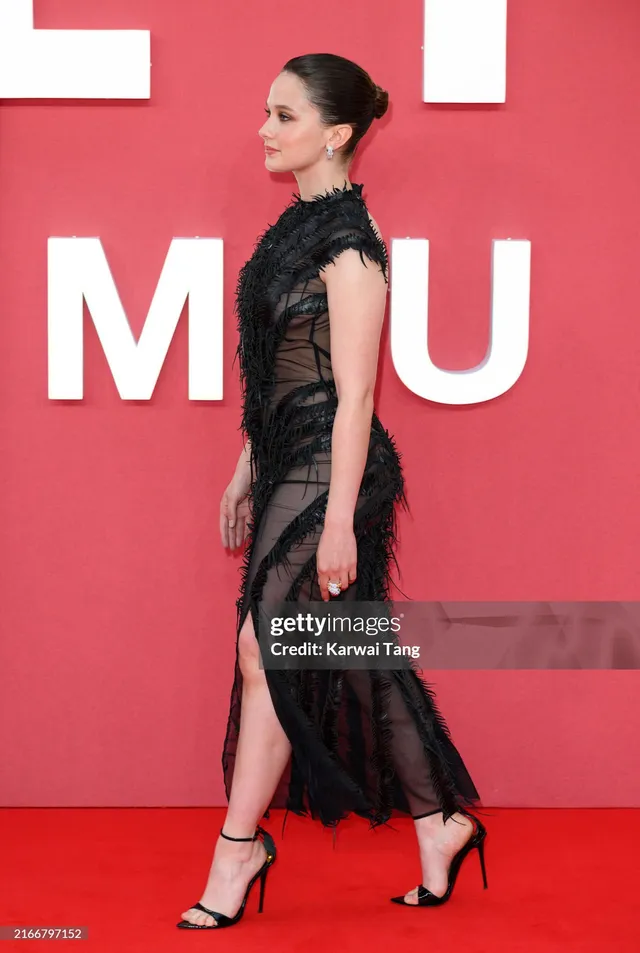 Cailee Spaeny wearing a black lace dress and black heels on a red carpet.