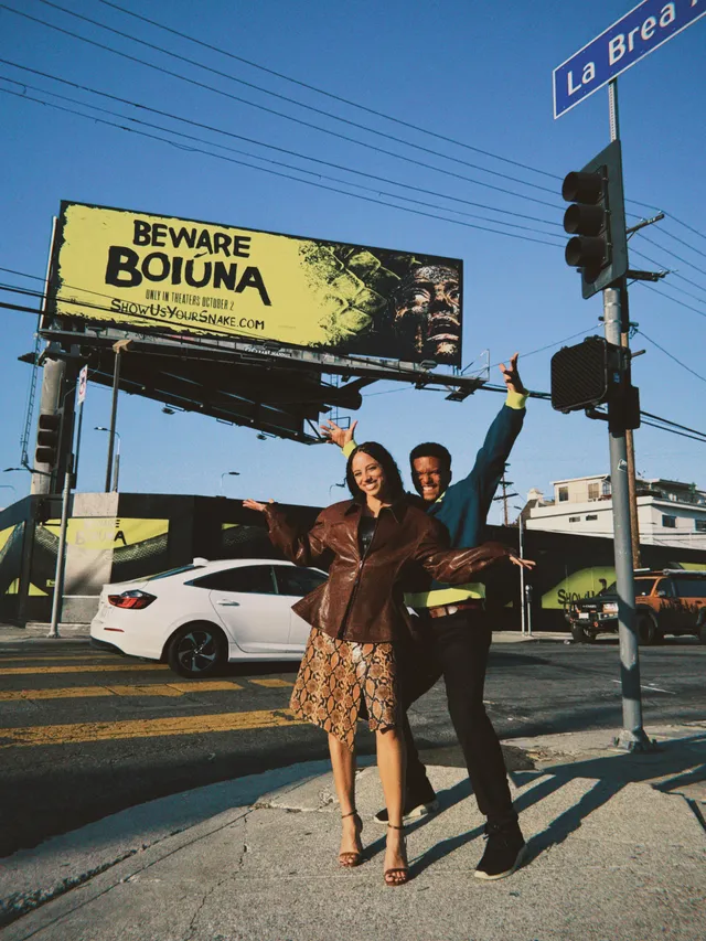 Kiana Madeira posing with a companion outdoors in a patterned dress and brown heels.