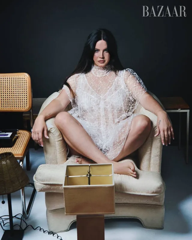 Lana Del Rey sitting cross-legged in an armchair in a lace dress.