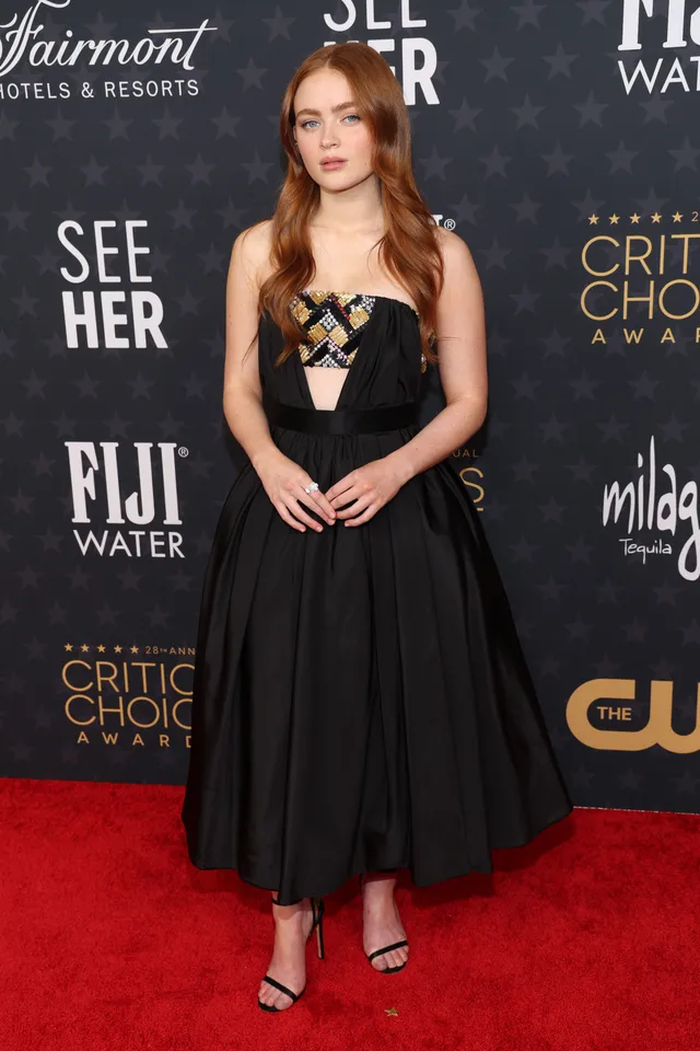 Sadie Sink standing in a black dress and strappy heels at a film event.