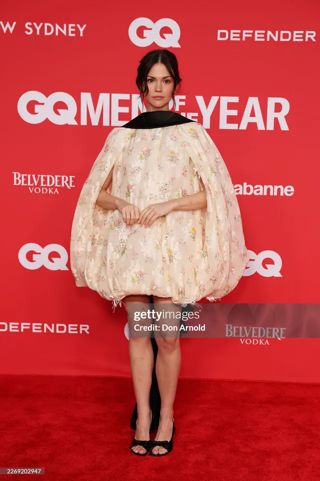 Maia Mitchell wearing a pale cape dress and black heels at an event.