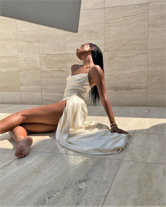 Jasmine Tookes sitting barefoot on a tiled floor in a pale dress.