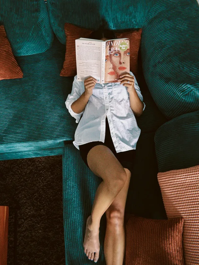 Kiana Madeira reclining barefoot on a green sofa while holding a book.