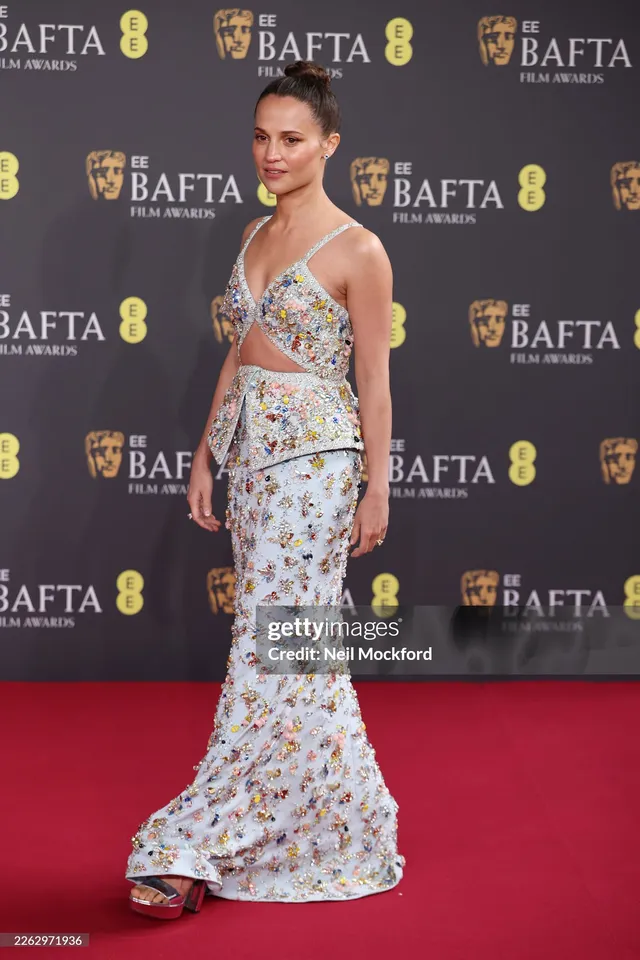 Alicia Vikander standing at a television event in a floral dress.