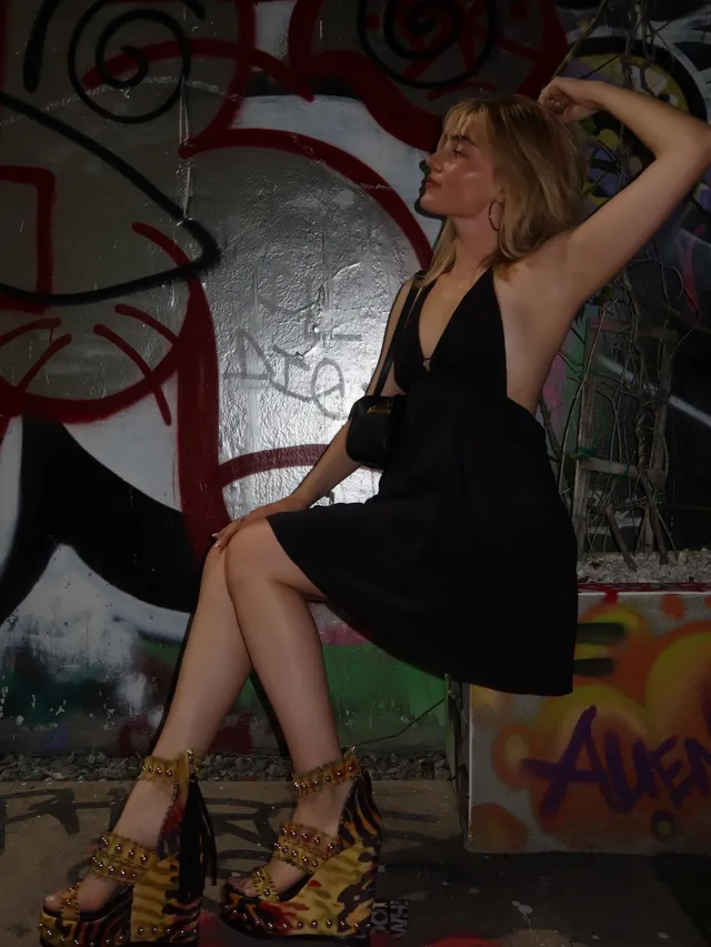 Meg Donnelly sitting by a graffiti wall in a black dress and patterned heels.