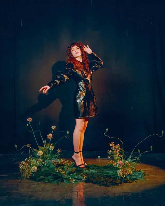 Sophie Thatcher posing on a decorated platform in a dark dress and heels.