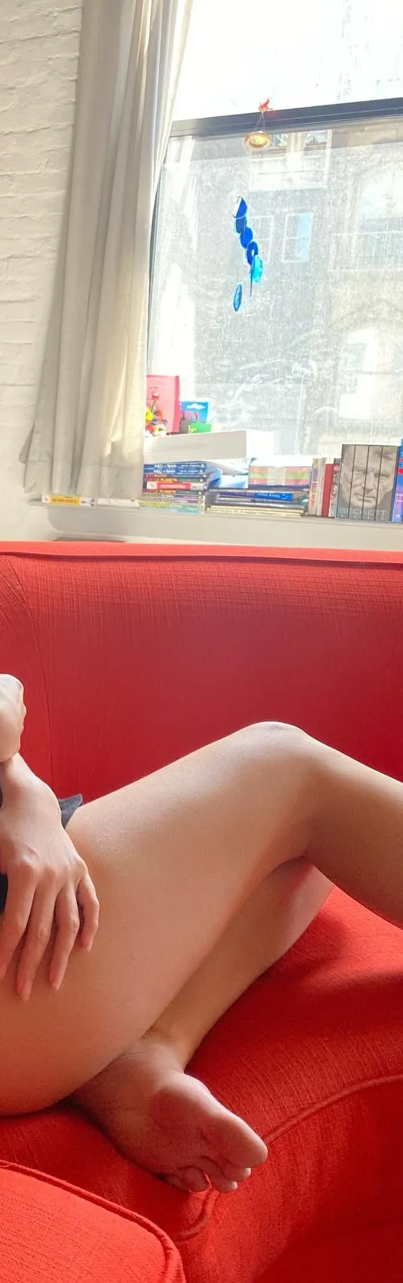 Emily Ratajkowski sitting barefoot on a red sofa.