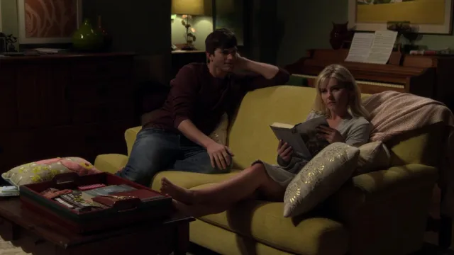 Elisha Cuthbert reclining barefoot on a sofa beside another person in a television scene.