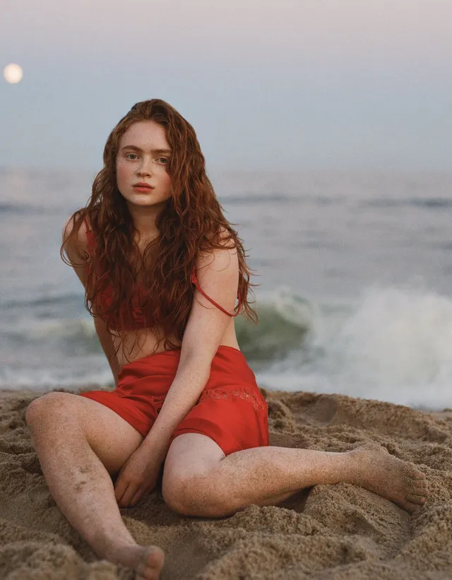 Sadie Sink sitting barefoot on sand in a red dress.