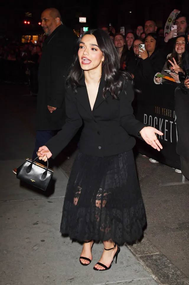 Rachel Zegler walking outside in a black lace dress and heeled sandals.